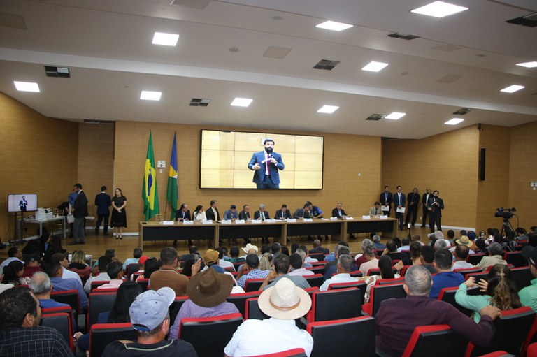 Foto mostra auditório da Assembleia Legislativa com pessoas sentadas, de costas, algumas com chapéus de boiadeiro. Ao fundo, uma mesa com autoridades e no telão a imagem do orador naquele momento, o procurador Gabriel de Amorim.