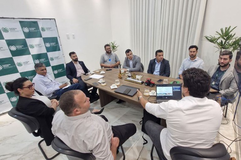 Foto mostra participantes da reunião, sentados em volta de uma mesa numa sala com paredes brancas e um banner em tons de verde e branco.