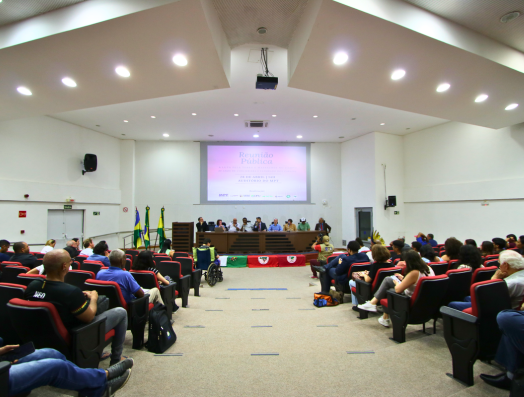 Público sentado em poltronas do auditório do Ministério Público do trabalho, acompanhando evento com mesa de autoridades ao fundo. Ao centro, há painel de projeção exibindo apresentação, e bandeiras estão posicionadas ao lado da mesa principal.
