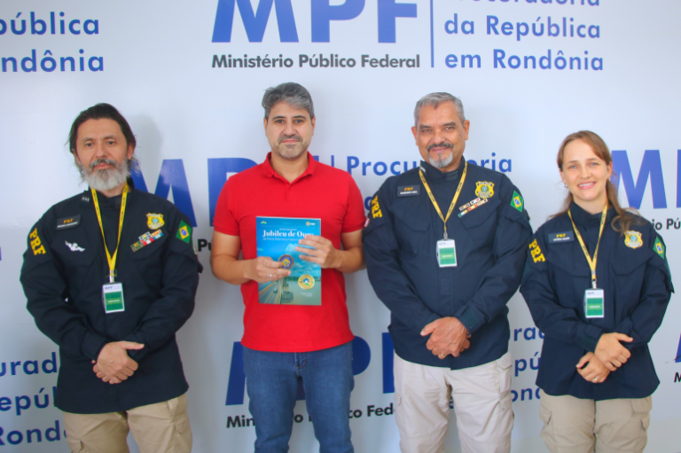 Fotografia de quatro pessoas posando lado a lado em frente a um painel com a identidade visual do Ministério Público Federal em Rondônia. Três delas usam uniformes azul-escuros com emblemas, bandeiras do Brasil e crachás pendurados no pescoço. A segunda pessoa da esquerda para a direita veste camiseta vermelha e segura um livro com capa azul e verde. Todos estão voltados para a câmera.