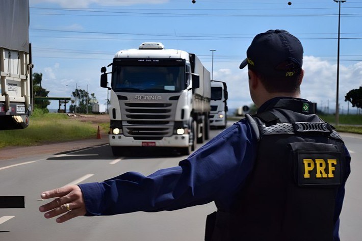 A imagem retrata uma fiscalização da Polícia Rodoviária Federal (PRF) em uma rodovia brasileira sob a luz do dia. Em primeiro plano, vemos um policial de costas, vestindo o uniforme azul-marinho com um colete tático ostentando a sigla "PRF" em letras amarelas, estendendo o braço esquerdo para sinalizar a parada de veículos. Ao fundo, um grande caminhão Scania branco se aproxima, seguido por outros caminhões de carga, indicando um ponto de controle de tráfego. O cenário é composto por uma pista asfáltica bem iluminada, vegetação nas laterais e um céu azul com poucas nuvens, transmitindo a ideia de uma operação de rotina para garantir a segurança viária ou fiscalizar o excesso de peso.
