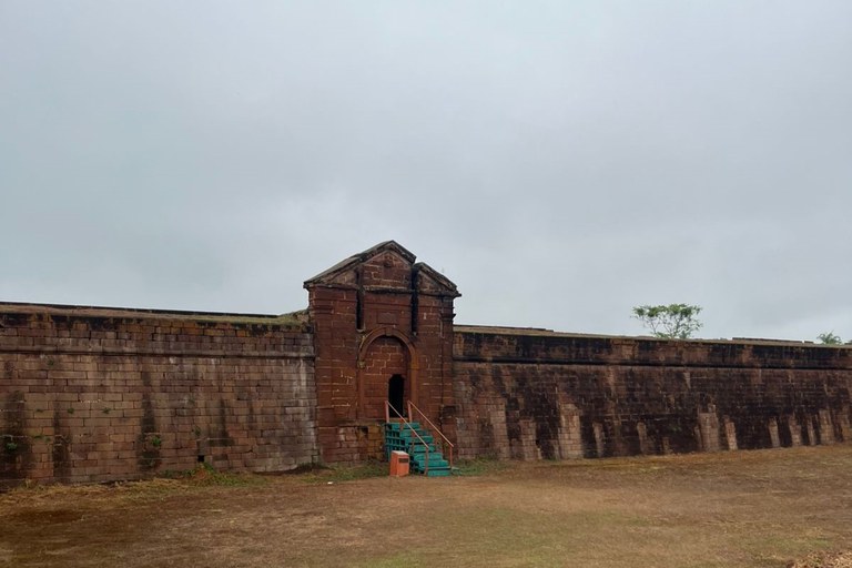 Foto em plano aberto mostra parte da muralha do Real Forte Príncipe da Beira, feita em pedra avermelhada. No centro, há um portão de entrada com uma pequena escada verde metálica. À frente, um amplo espaço de chão de terra e gramado ralo. O céu está nublado, dando aspecto cinzento à cena.