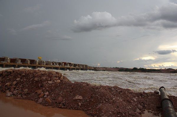 Uma imagem horizontal mostra a Usina Hidrelétrica de Jirau sob um céu carregado de nuvens escuras e cinzentas. Ao fundo, no centro e à esquerda, estende-se a longa estrutura de concreto da barragem, de onde a água flui intensamente, criando uma forte correnteza de tom barrento que ocupa a metade direita da cena. No primeiro plano, à esquerda, há um grande monte de terra e pedras avermelhadas, ladeado por uma poça de água marrom. No canto inferior direito, um longo tubo metálico escuro avança em direção à água. O horizonte distante exibe uma faixa de vegetação baixa sob um vislumbre de claridade no fim da tarde.