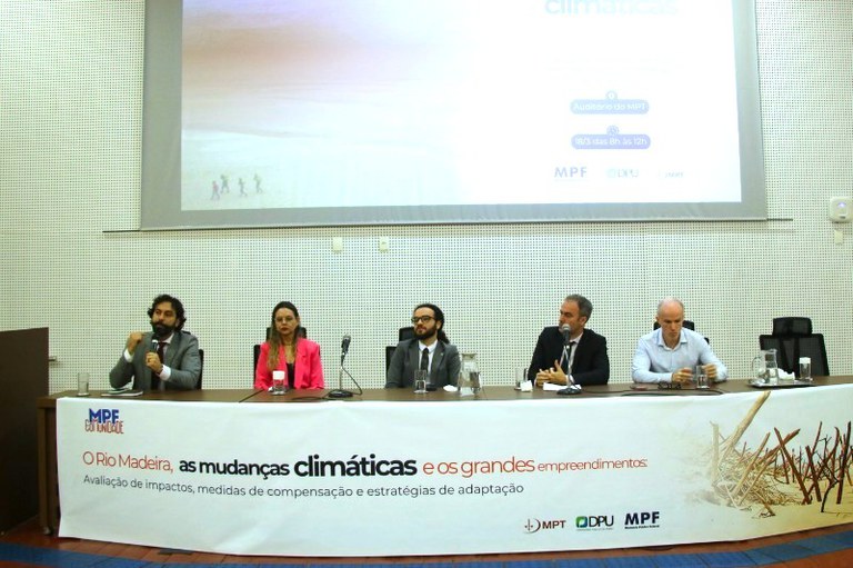 A imagem registra a composição de uma mesa redonda durante um evento institucional, onde cinco especialistas estão sentados lado a lado em um auditório. À frente deles, um banner branco estendido na mesa exibe o título do debate em português: "O Rio Madeira, as mudanças climáticas e os grandes empreendimentos", acompanhado pelos logotipos do Ministério Público Federal (MPF), da Defensoria Pública da União (DPU) e do Ministério Público do Trabalho (MPT). Ao fundo, uma grande tela de projeção repete as informações do evento, indicando que a discussão foca na avaliação de impactos e estratégias de adaptação. O ambiente possui um tom formal, com paredes acústicas claras, e os participantes — quatro homens e uma mulher — aparecem em meio a falas e anotações, sugerindo um momento de intercâmbio técnico e jurídico sobre questões ambientais na Amazônia.