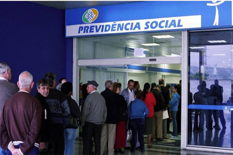 Fotografia mostra uma fila de pessoas em frente a uma agência da Previdência Social, com a placa “Previdência Social” acima da entrada.