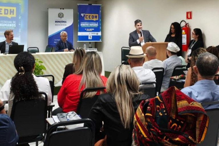 Foto de um evento. Há pessoas sentadas de costas. Na frente, em um púlpito, em pé, do lado direito da foto, o procurador regional dos Direitos do Cidadão (PRDC), Raphael Luís Pereira Bevilaqua faz pronunciamento. Do lado esquerdo, é possível ver uma mesa com duas pessoas.