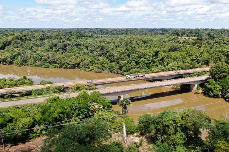 A imagem mostra uma vista aérea de uma ponte sobre um rio, cercada por uma floresta densa. A ponte tem veículos, incluindo um caminhão e carros, atravessando-a.
