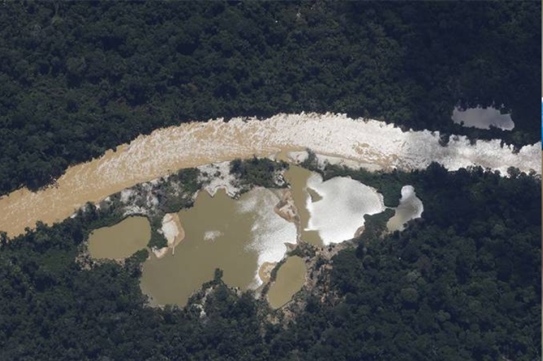 Esta fotografia aérea captura uma área de severo impacto ambiental em Vilhena, Rondônia, dividida nitidamente por uma estrada de terra central que corta a paisagem de forma vertical. À esquerda da via, destaca-se uma imensa cratera de mineração ou garimpo com solo arenoso e amarelado totalmente exposto, cercada por altos barrancos escavados e com um pequeno caminhão estacionado próximo à entrada. À direita da estrada, estende-se um vasto terreno plano marcado pelo desmatamento recente e por uma área queimada, coberta por cinzas escuras e marcas de maquinário. Ao fundo e nas extremidades, faixas remanescentes de floresta verde e vegetação rasteira resistem, evidenciando o forte contraste entre a natureza preservada e a destruição provocada pela ação humana.