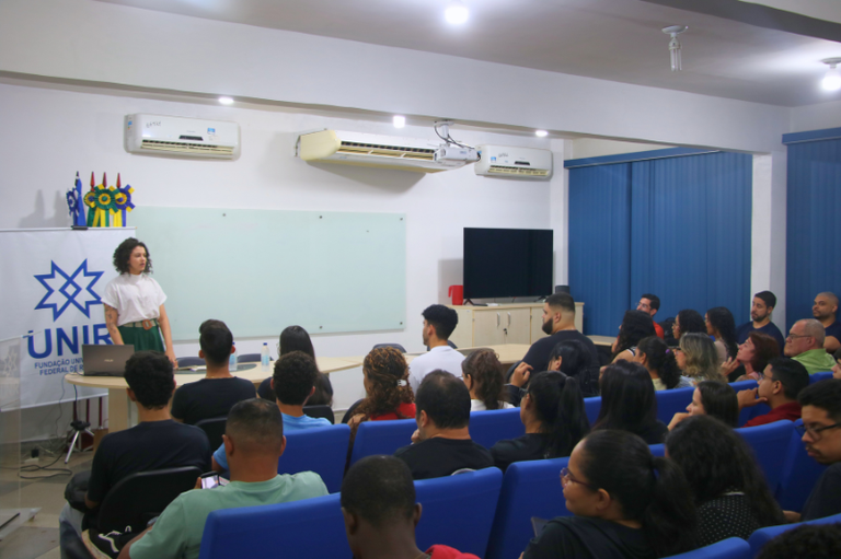 Uma mulher de cabelos cacheados apresenta uma palestra para um auditório atento na Universidade Federal de Rondônia (UNIR). À esquerda, há um banner com o logotipo da instituição; ao fundo, uma lousa de vidro e equipamentos de ar-condicionado.