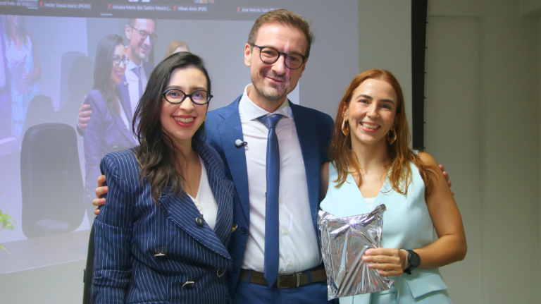 Fotografia de três pessoas sorrindo e posando lado a lado em um ambiente interno. Ao centro, um homem usa terno azul, camisa branca, gravata e óculos. À esquerda, uma mulher usa óculos e um conjunto social azul com listras. À direita, uma mulher usa um conjunto azul-claro e segura um objeto embrulhado em papel prateado. Ao fundo, há uma tela de projeção com imagens de um evento.