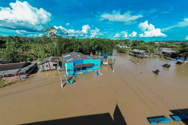 Fotografia de uma rua alagada, com várias casas e estabelecimentos parcialmente submersos pelas cheias do rio Maderia, em 2025. Algumas das casas são azuis e há densa vegetação ao fundo