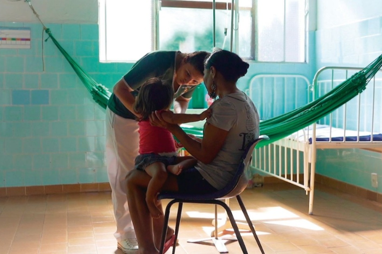 Em um ambiente hospitalar, com paredes azul-claro e camas de metal, uma mulher sentada segura uma criança pequena no colo, enquanto uma profissional de saúde interage com a criança, olhando atentamente para ela. Ao fundo, há uma rede verde pendurada entre duas paredes e uma cama hospitalar vazia. A cena é iluminada por luz natural que entra pela janela, transmitindo acolhimento e cuidado.