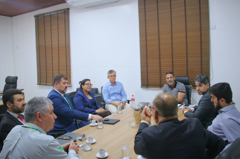 Nove pessoas, majoritariamente homens em trajes formais e uma mulher, participam de uma reunião de negócios ou institucional em torno de uma grande mesa de conferência de madeira. Há copos d'água e xícaras de café na mesa.
