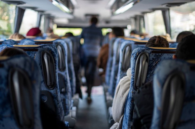 Foto mostra o interior de um ônibus com as poltronas e pessoas sentadas. No corredor há uma pessoa em pé