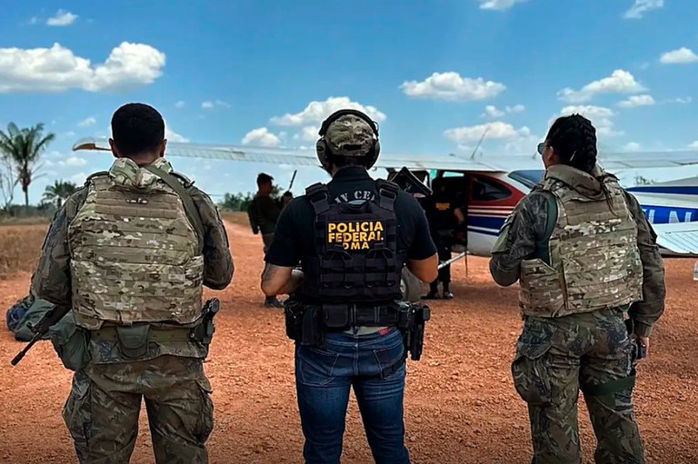 A foto, tirada em uma área de solo avermelhado, possivelmente no contexto de uma operação de segurança ou fiscalização, mostra três pessoas em primeiro plano, vistas de costas, que parecem ser agentes de segurança ou militares devido aos seus equipamentos e vestimentas. O indivíduo central veste uma camiseta preta e calças jeans, com um colete tático preto onde se lê "POLÍCIA FEDERAL DO MA", além de um boné. Ele está flanqueado por duas pessoas vestindo coletes táticos e uniformes camuflados. Ao fundo, um avião monomotor de pequeno porte, com detalhes em vermelho e branco, está estacionado, e outras figuras podem ser vistas perto ou dentro da aeronave, sugerindo a intercepção ou inspeção da mesma. O cenário é de mata rasteira ou campo aberto, sob um céu claro com algumas nuvens.