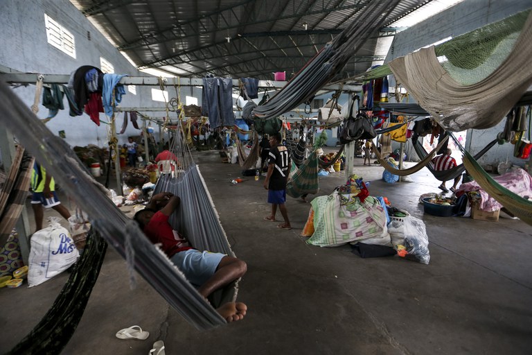 A foto mostra o abrigo Janokoida, em Pacaraima, o ambiente é amplo e coberto, com iluminação natural entrando por janelas altas nas paredes laterais. Diversas redes estão penduradas em estruturas metálicas, sendo utilizadas por pessoas para descanso ou acomodação. Há itens pessoais, como roupas, sacolas e objetos espalhados pelo local, organizados em torno das redes. Algumas pessoas estão deitadas, enquanto outras estão em pé ou realizando atividades. O teto é de estrutura metálica, e o piso é de concreto.