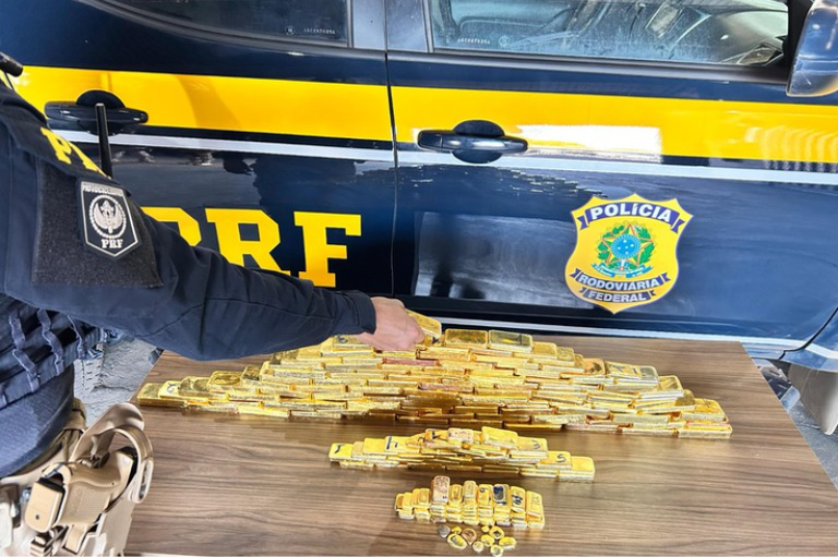 Foto mostra dezenas de barras de ouro empilhadas sobre uma mesa em frente a uma viatura da Polícia Rodoviária Federal (PRF), com um policial fardado ao lado.