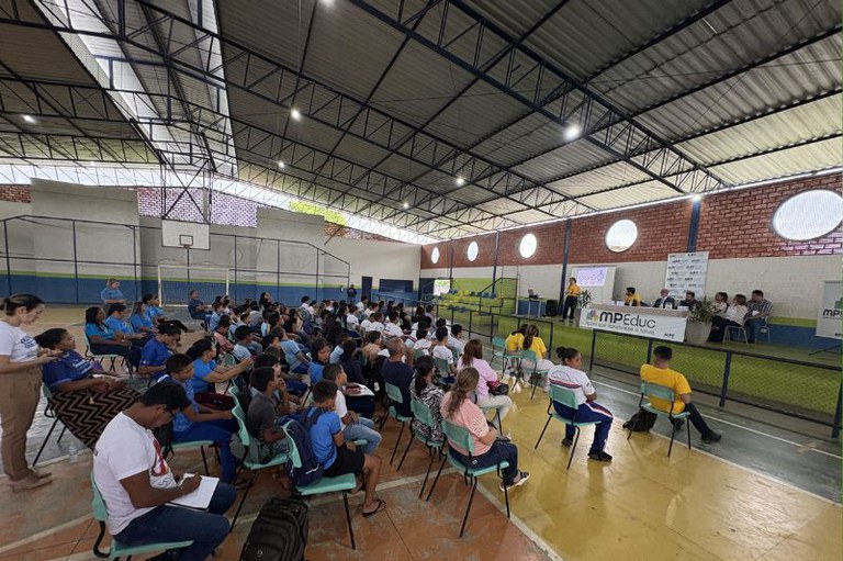 Foto mostra o evento em um ginásio com as pessoas sentadas em cadeiras e a mesa com os palestrantes