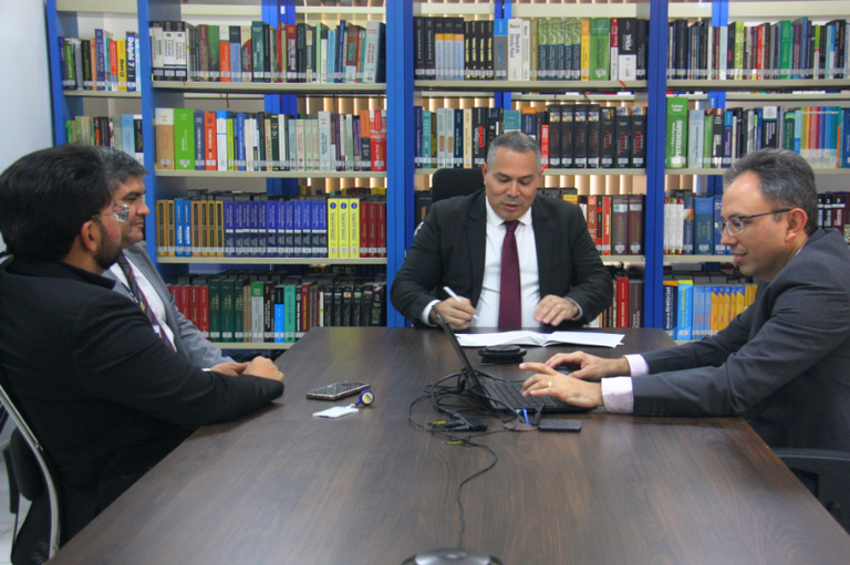 Quatro homens de terno estão reunidos em torno de uma mesa retangular. Um homem assina papéis e outro utiliza um computador. No fundo, aparecem estantes cheias de livros organizados. Sobre a mesa, existem dispositivos eletrônicos, como um telefone, cabos e um gravador.