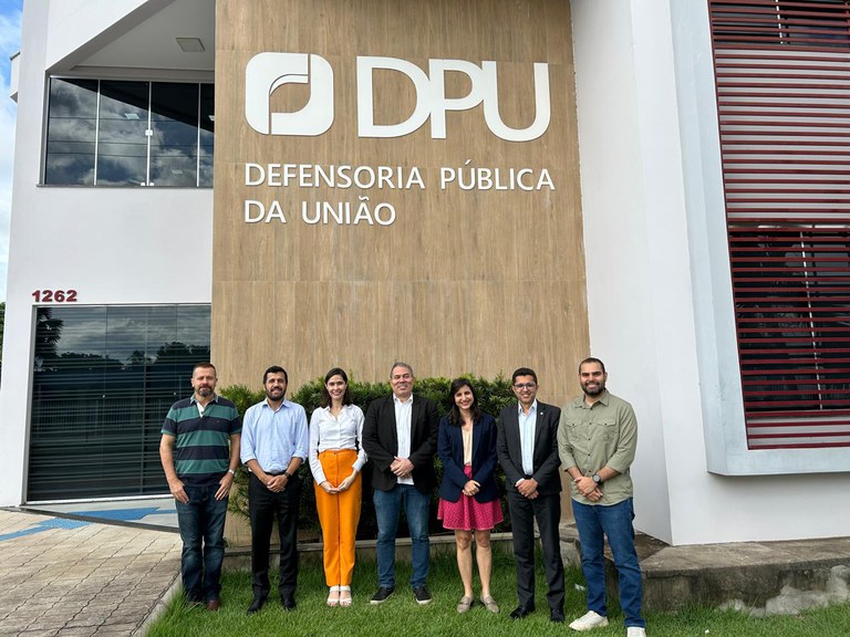 Fotografia mostra sete pessoas (sendo duas mulheres e cinco homens) posando, lado a lado, em frente à sede da Defensoria Pública da União em Roraima