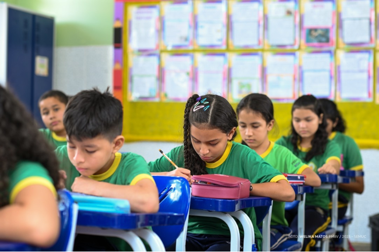 Foto mostra crianças em uniformes verdes sentadas em carteiras azuis em uma sala de aula, concentradas em escrever em seus cadernos.