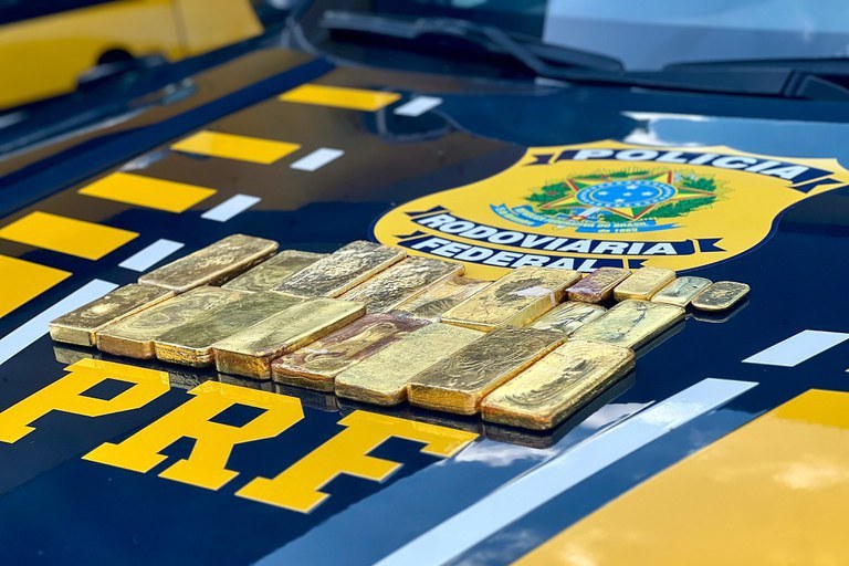 A imagem mostra diversas barras de ouro de diferentes tamanhos e formatos irregulares dispostas sobre o capô de uma viatura da Polícia Rodoviária Federal (PRF) do Brasil. O veículo é preto com detalhes em amarelo vibrante, exibindo o logotipo oficial da corporação ao fundo e a sigla "PRF" em destaque no primeiro plano. O ouro apresenta uma textura rústica, sugerindo ser fruto de fundição artesanal ou garimpo, e a composição da foto enfatiza o resultado de uma apreensão policial realizada em ambiente externo sob luz natural.