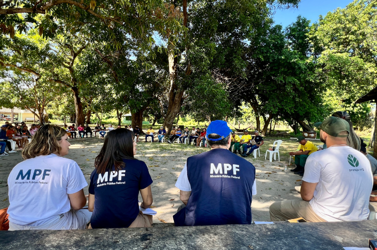 Integrantes do Ministério Público Federal (MPF), vistos de costas em primeiro plano, participam de uma reunião ao ar livre com um grupo de pessoas dispostas em círculo sob a sombra de grandes árvores.