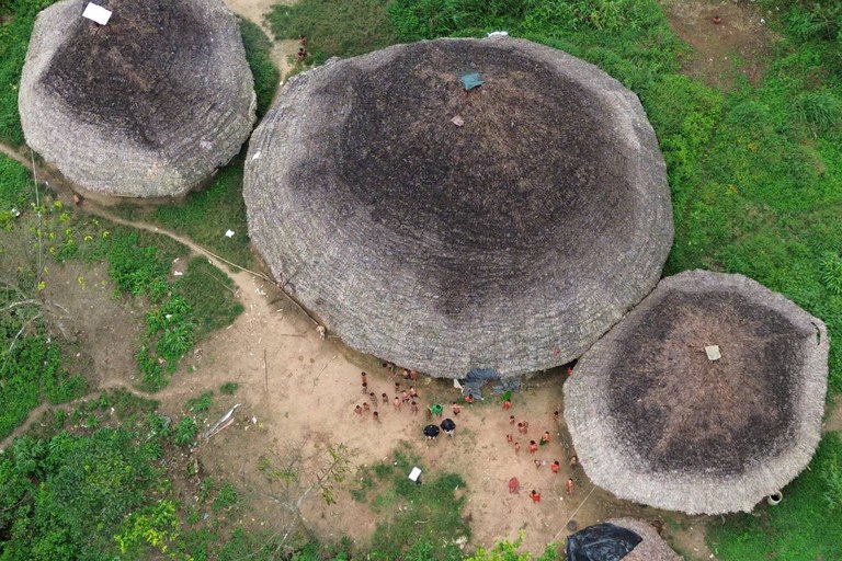 Fotografia aérea com foco em três grandes ocas tradicionais com telhados arredondados de palha em tons de cinza e marrom, dispostas sobre um terreno de terra batida cercado por vegetação verde densa. No centro, sob o maior telhado de palha, dezenas de pessoas indígenas e alguns visitantes estão reunidos ao redor de uma pequena área limpa no chão. Um caminho de terra estreito serpenteia pelo canto esquerdo, conectando as estruturas em meio ao ambiente natural.