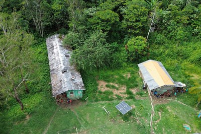 Uma vista aérea mostra um pequeno povoado aninhado em uma floresta tropical verdejante. Duas construções de madeira com telhados de palha e zinco estão dispostas no centro, cercadas por vegetação e um rio estreito que serpenteia pelo lado esquerdo da imagem. As estruturas são ligadas por passarelas de madeira que atravessam a água e o terreno irregular. Uma área de terra batida se estende na frente das casas, com uma pequena horta e árvores frutíferas próximas. No topo da maior casa, um pequeno barco com cobertura está atracado na margem do rio.