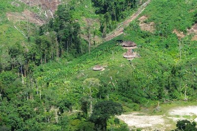 Fotografia aérea panorâmica mostra uma comunidade indígena em uma encosta íngreme cercada por mata densa e áreas com vegetação rasteira e clareiras de desmatamento. Ao longo do morro, sobressaem-se três ocas tradicionais redondas com telhados de palha, dispostas em diferentes níveis entre plantações e pequenas clareiras. No canto inferior direito, é visível um trecho de solo exposto de areia clara ao lado da vegetação.
