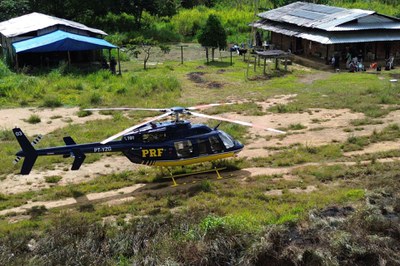 Um helicóptero da Polícia Rodoviária Federal (PRF), pintado de azul e amarelo com a matrícula PT-YZG e o prefixo 191 visíveis na cauda, está pousado em uma clareira de grama alta e solo exposto em uma área rural. Ao fundo, à direita, há uma grande casa de madeira e telhado de zinco, sob a qual algumas pessoas parecem estar reunidas ou trabalhando. À esquerda, uma estrutura de madeira menor e mais simples é parcialmente coberta por uma lona azul, localizada sob uma grande árvore. A área é cercada por vegetação nativa densa. A luz solar é intensa e projeta sombras fortes sobre o solo.