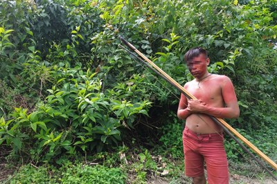 Um homem indígena está em pé diante de uma densa vegetação folhosa, segurando um grande arco e flechas artesanais na posição diagonal. Ele está sem camisa, vestindo apenas um short vermelho, e apresenta pintura corporal avermelhada no tórax, braços e rosto, que tem a área dos olhos pixelada para preservar sua identidade.