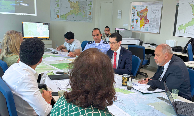 A foto mostra uma reunião em uma sala com várias pessoas sentadas ao redor de uma mesa grande, sobre a qual há mapas e documentos espalhados. O procurador Andre Luiz Porraca está ao centro, usando terno e gravata vermelha, com outros participantes ao redor. Um monitor ao fundo exibe um mapa, e as paredes também têm mapas de Roraima fixados. A iluminação é clara, e a sala possui cadeiras azuis e móveis em tons neutros.