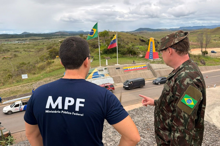 Foto mostra um integrante do MPF e um militar fardado observando o posto de controle na fronteira entre Brasil e Venezuela, sinalizado por bandeiras e um marco divisório