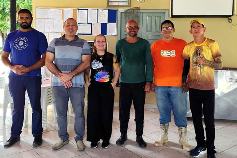 Seis pessoas estão em pé, lado a lado, posando para a foto em um ambiente interno. Atrás delas há uma parede amarela com murais de avisos, porta, quadro branco, relógio e algumas fotos em moldura.