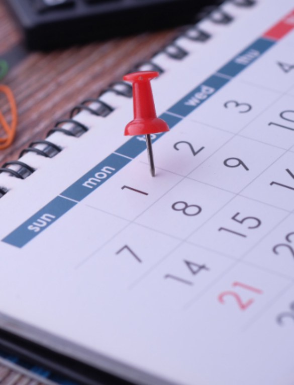 A imagem mostra um calendário de mesa com um alfinete vermelho marcando o dia 1, que é uma segunda-feira.O número 21 está destacado em vermelho, sugerindo uma data especial ou feriado. No fundo, desfocados, há alguns objetos de escritório como um clipe laranja e parte de uma calculadora.