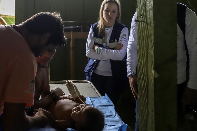 Foto mostra bebê yanomami em atendimento de saúde, deitado sobre maca, em área interna de unidade de saúde. Ao lado da maca, há dois homens segurando as mãos do bebê. Ao fundo, há uma mulher loira e um homem em pé, com uniforme da Sesai