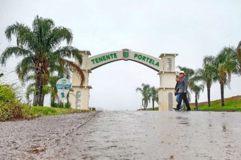 Pórtico de entrada com a inscrição "TENENTE PORTELA", ladeado por palmeiras, com duas pessoas caminhando ao lado de uma via molhada.