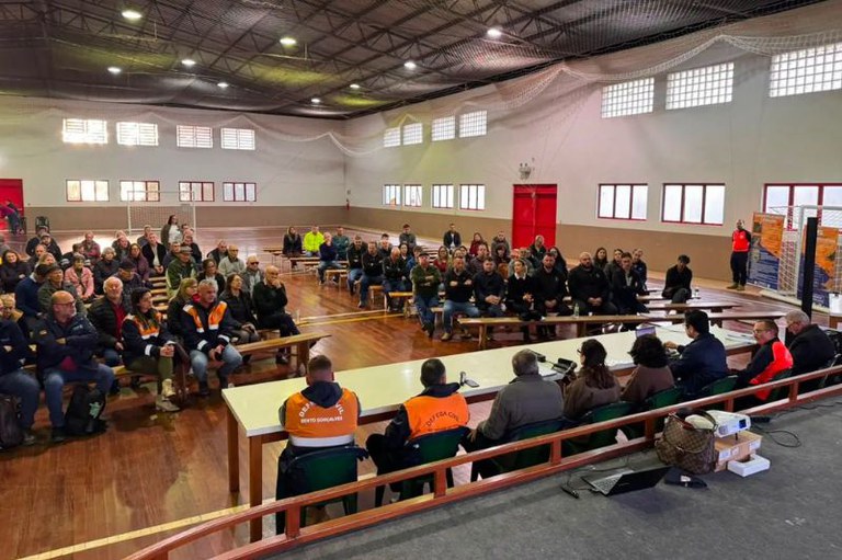 Foto mostra salão onde foi realizada a audiência com o público sentado em bancos de frente para a mesa com 8 palestrantes, que estão de costas