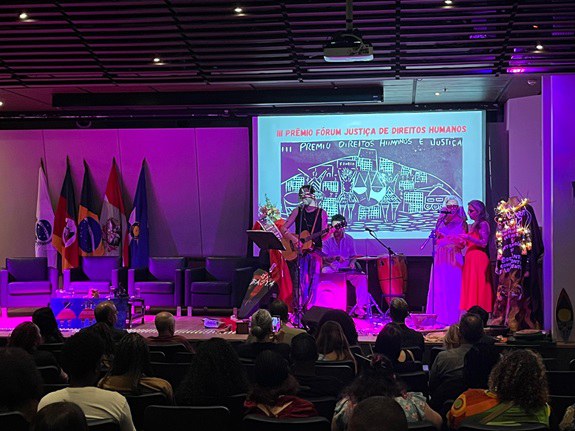 Foto mostra auditório com pessoas sentadas de costas e ao fundo a banda cultura se apresentando no palco