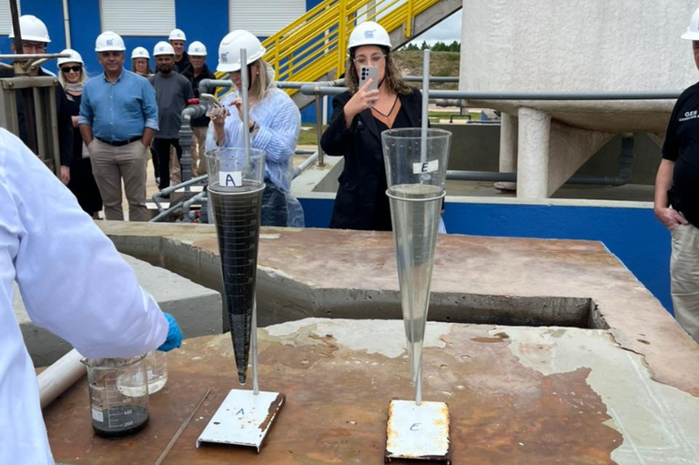 Inspeção ETE Cidreira Dois cones de sedimentação (cones Imhoff) posicionados lado a lado em uma base de concreto, comparando amostras de água de uma estação de tratamento. O cone à esquerda contém um líquido escuro e denso, enquanto o cone à direita apresenta um líquido claro e límpido. Ao fundo, um grupo de profissionais com capacetes brancos observa e fotografa o experimento. No canto esquerdo, parte do braço de um técnico em jaleco branco e luvas azuis manipula béqueres de laboratório.