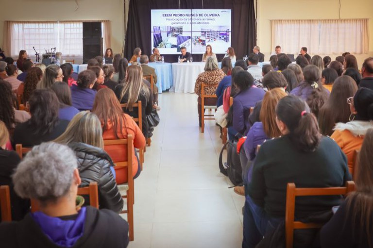Vista traseira de uma plateia lotada assistindo a uma apresentação. Ao fundo, palestrantes estão reunidos em uma mesa diante de uma tela que exibe o título "EEEM Pedro Nunes de Oliveira" e fotos de um projeto de acessibilidade.