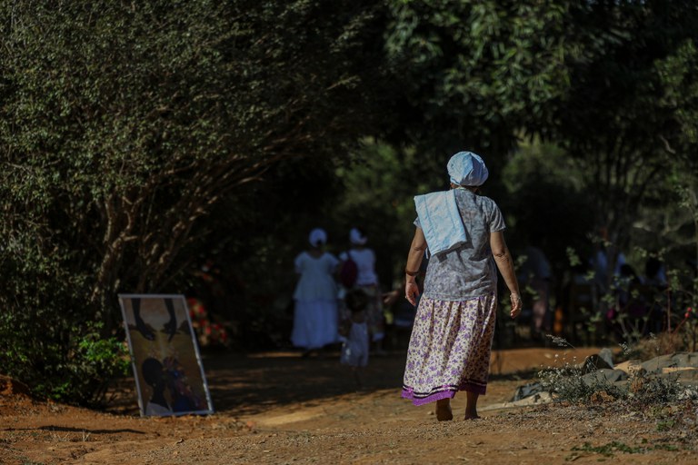 Foto mostra uma mulher idosa com lenço branco na cabeça caminhando em estrada de terra em direção a um grupo de pessoas de roupas brancas, em uma comunidade tradicional ou quilombo.