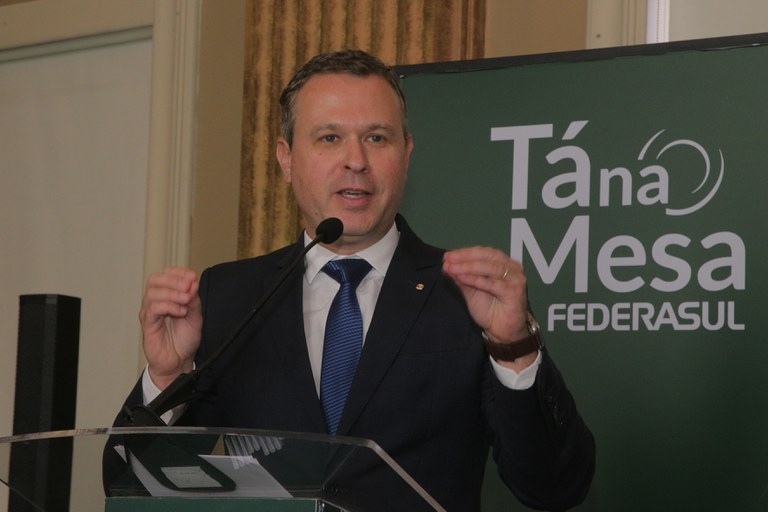 Procurador-chefe falando ao microfone, de terno e com as mãos erguidas, tendo ao fundo o painel verde com o logo do evento Tá na Mesa da Federasul