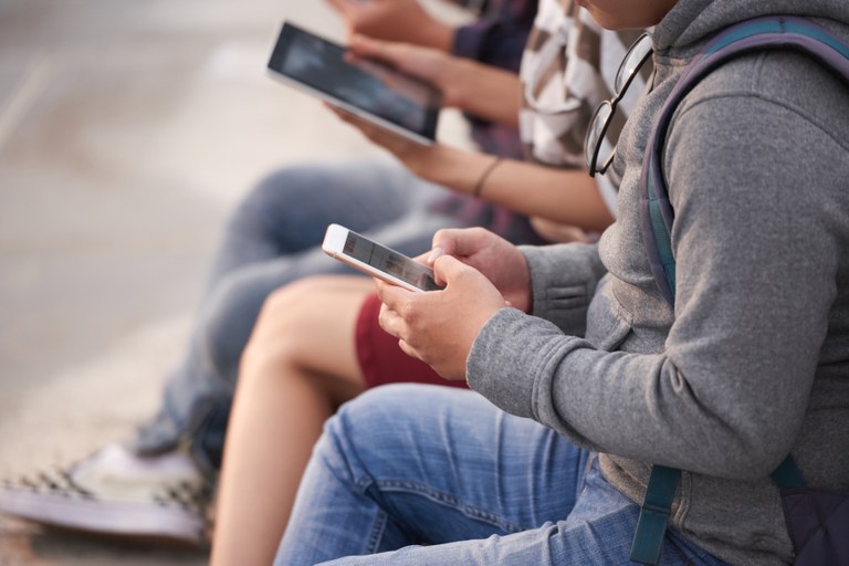 Grupo de jovens sentados lado a lado ao ar livre, concentrados no uso de celulares e tablet. Em primeiro plano, uma pessoa vestindo casaco cinza com capuz e mochila segura um smartphone com ambas as mãos, enquanto as pessoas ao lado utilizam outros dispositivos móveis.