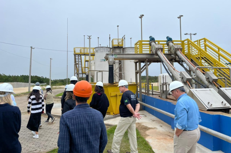 Um grupo de pessoas usando capacetes de segurança realiza uma inspeção técnica em uma estação de tratamento de efluentes (ETE). Em primeiro plano, os visitantes observam as instalações, que incluem tanques cilíndricos brancos, passarelas metálicas amarelas e grandes tubulações inclinadas para o transporte de resíduos. O ambiente é externo, sob um céu nublado, com pavimentação de blocos intertravados e gramado ao redor das estruturas.