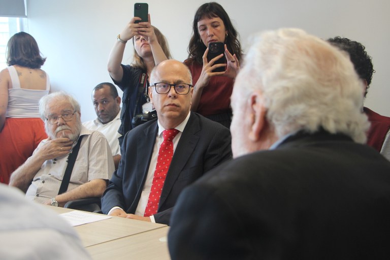Na imagem, o procurador da República Enrico de Freitas aparece sentado à mesa, observando o advogado Jair Krischke, que conversa com ele e aparece de costas. Ambos estão cercados de mais participantes da reunião