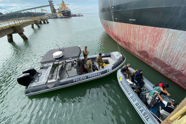 Agentes da Polícia Federal e da Marinha do Brasil em botes de patrulha ao lado de um navio de carga atracado em um porto.