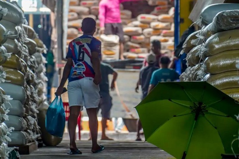 A foto mostra um corredor de armazém ladeado por sacos empilhados de comida. Em primeiro plano, um homem de costas carrega uma sacola em direção a uma área externa iluminada. Ao fundo, trabalhadores realizam o descarregamento de sacos de alimento em um caminhão.
