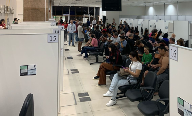 Imagem mostra pessoas sentadas numa sala com várias divisórias aguardando serem chamadas para o atendimento